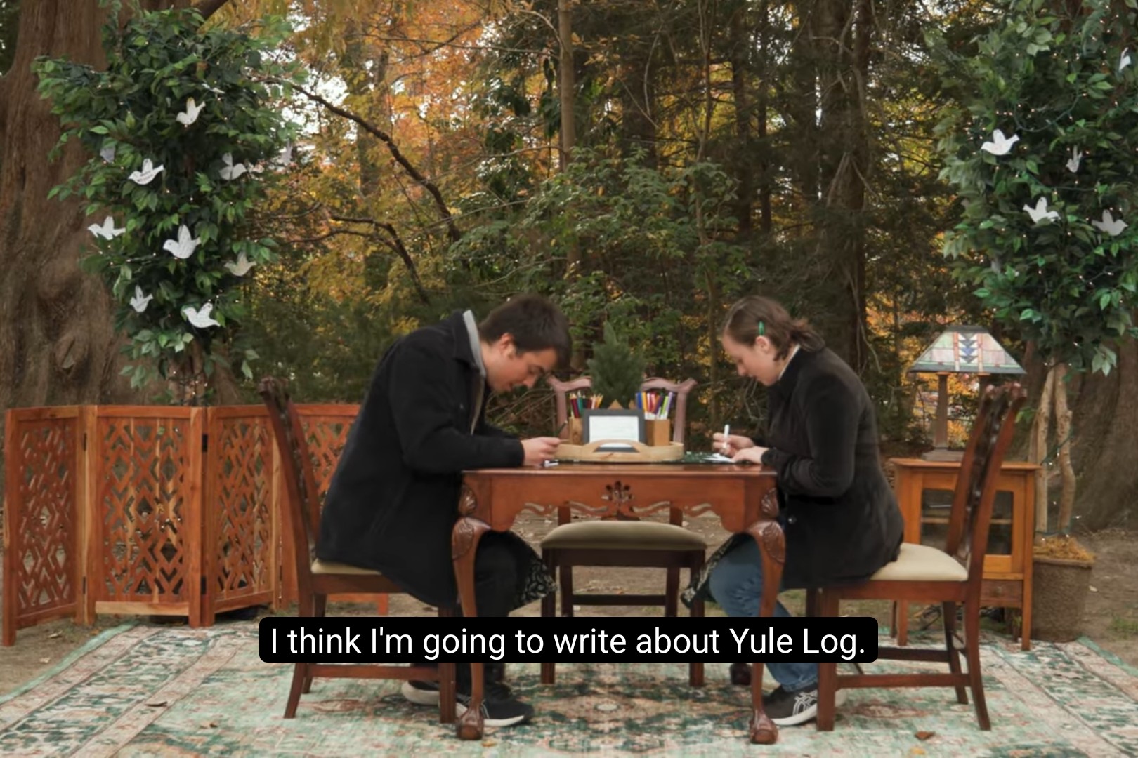 Two William & Mary students sit at an ornate table writing cards. One of them says, "I think I'm going to write about Yule Log."