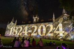A light show is displayed on the side of an older brick building. The lights display the years "1824-2024."