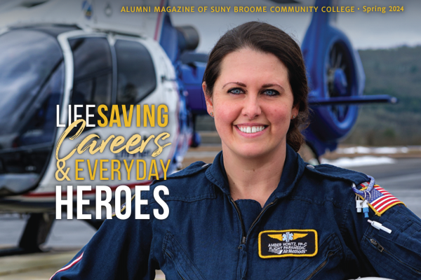 The cover of BROOME alumni magazine's spring 2024 issue shows a flight paramedic smiling for camera. She holds her flight helmet under one arm and a helicopter can be seen on a landing pad in the background.