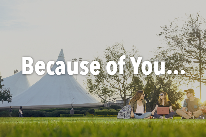 A photo of three students sitting together on a lawn. Behind them are trees and campus buildings.