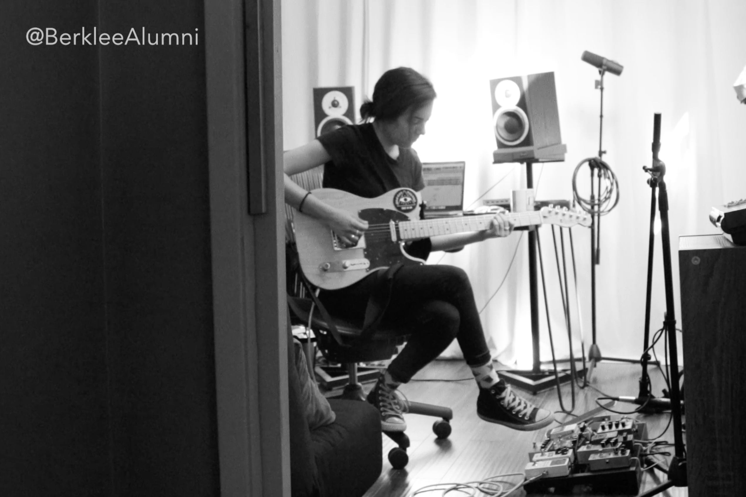 A black and white photo of a student playing guitar with studio equipment surrounding them.