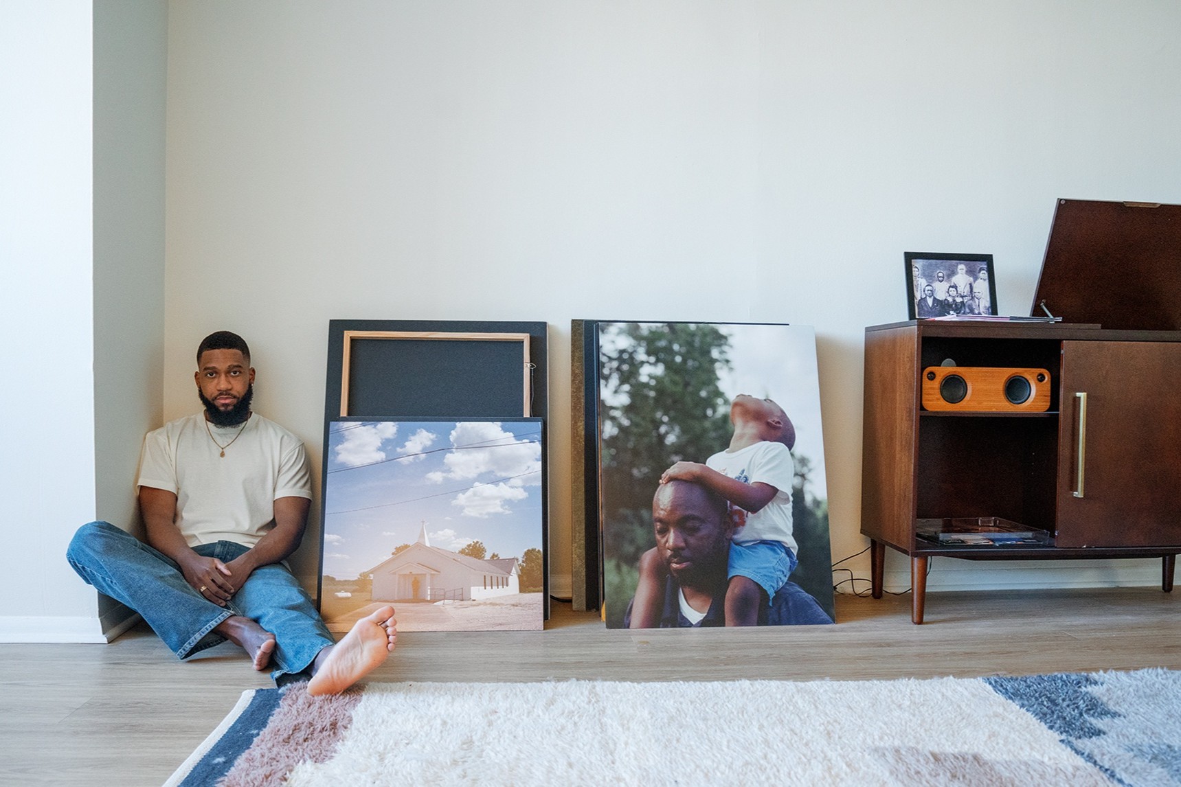 John Johnson sits on the floor next to prints of his own photography. Further along the wall is a wooden record stand and player.