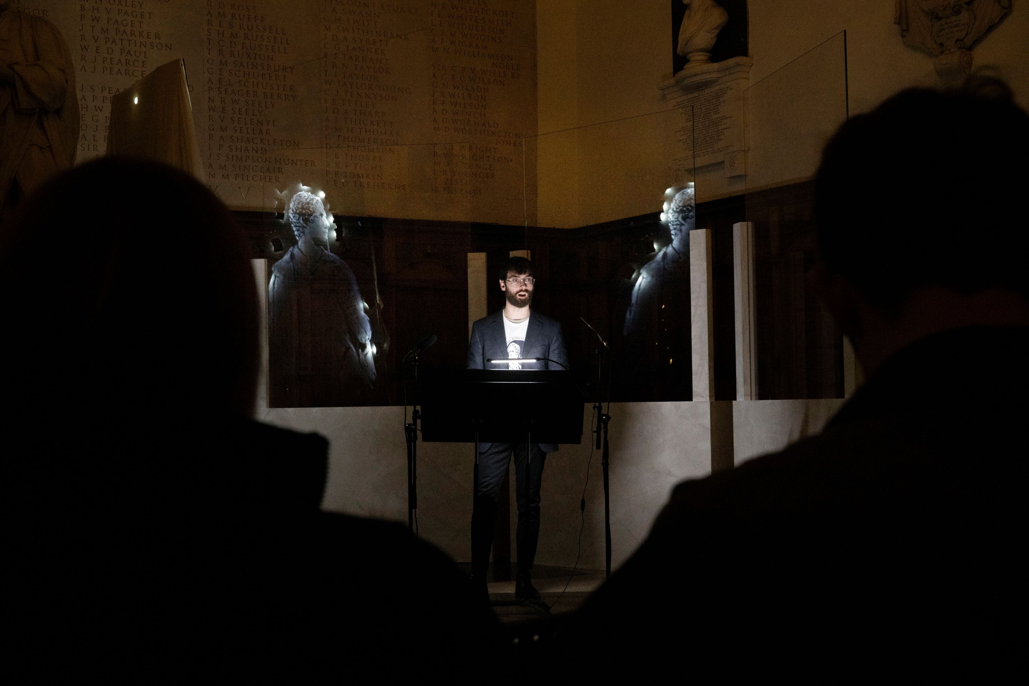 A man performs a reading in a darkened room, images of Byron projected behind him.