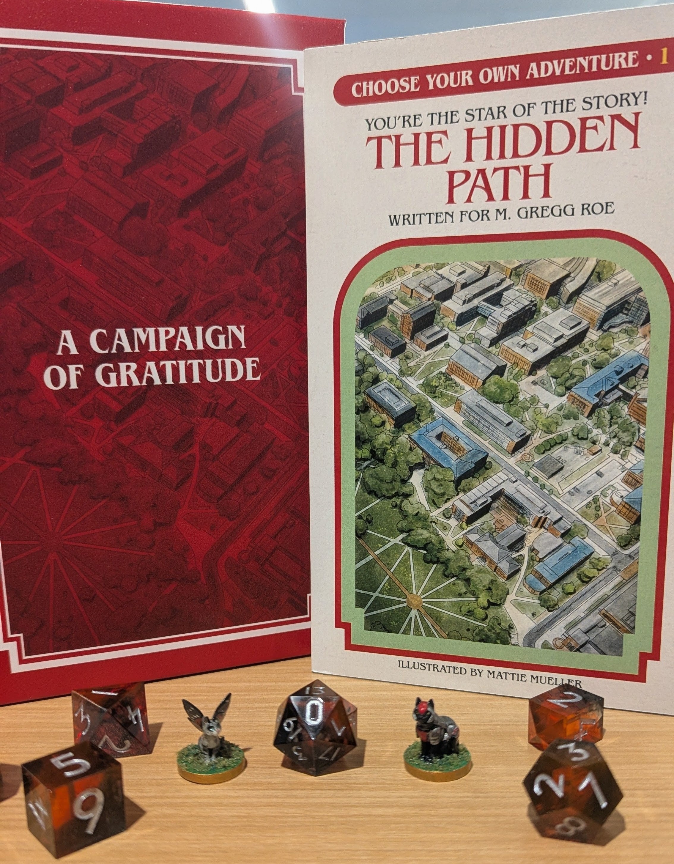 A photo of The Ohio State University's Choose Your Own Adventure style gift for a donor. The cover of the book reads, "The Hidden Path," and features an illustration of the campus. In front of the book on a table are D&D dice and two small cat figurines.