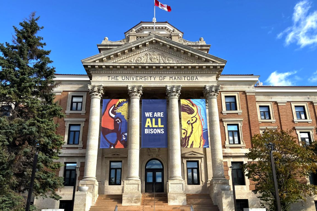 A photo of a columned building on the University of Manitoba campus. A series of three red, blue, and yellow banners hang between the building's columns. The right and left banner shows images of a bison, with the middle banner reading "We are all Bisons."