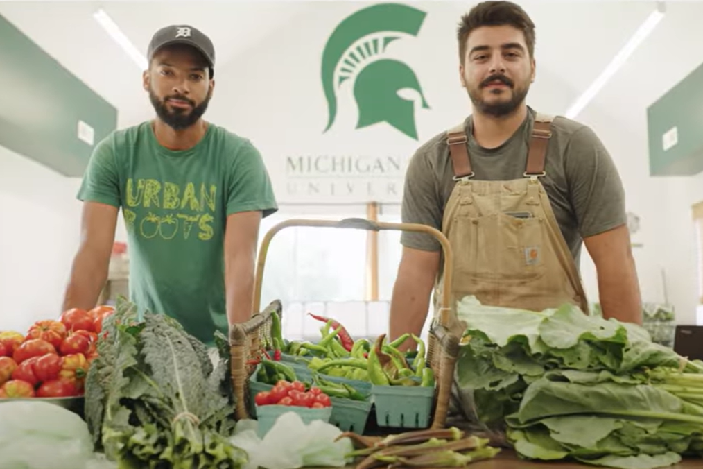 Two people wearing Michigan-green stand behind a table covered in fresh vegetables, including tomatoes, peppers, and leafy greens.
