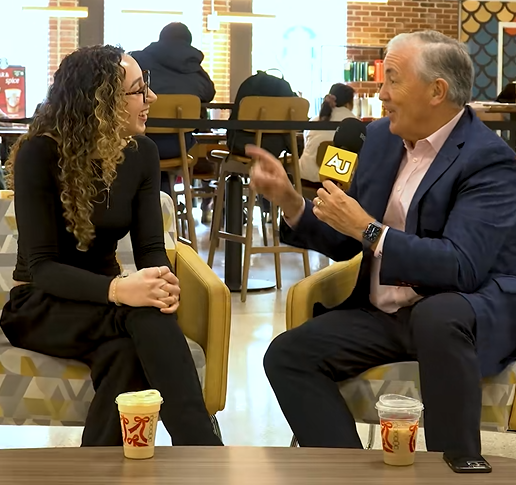 In a coffee shop, Board Chair Michael A. L. Balboni and Adelphi student Emily Munayirji sit in side by side chairs. Michael holds a microphone, interviewing her.
