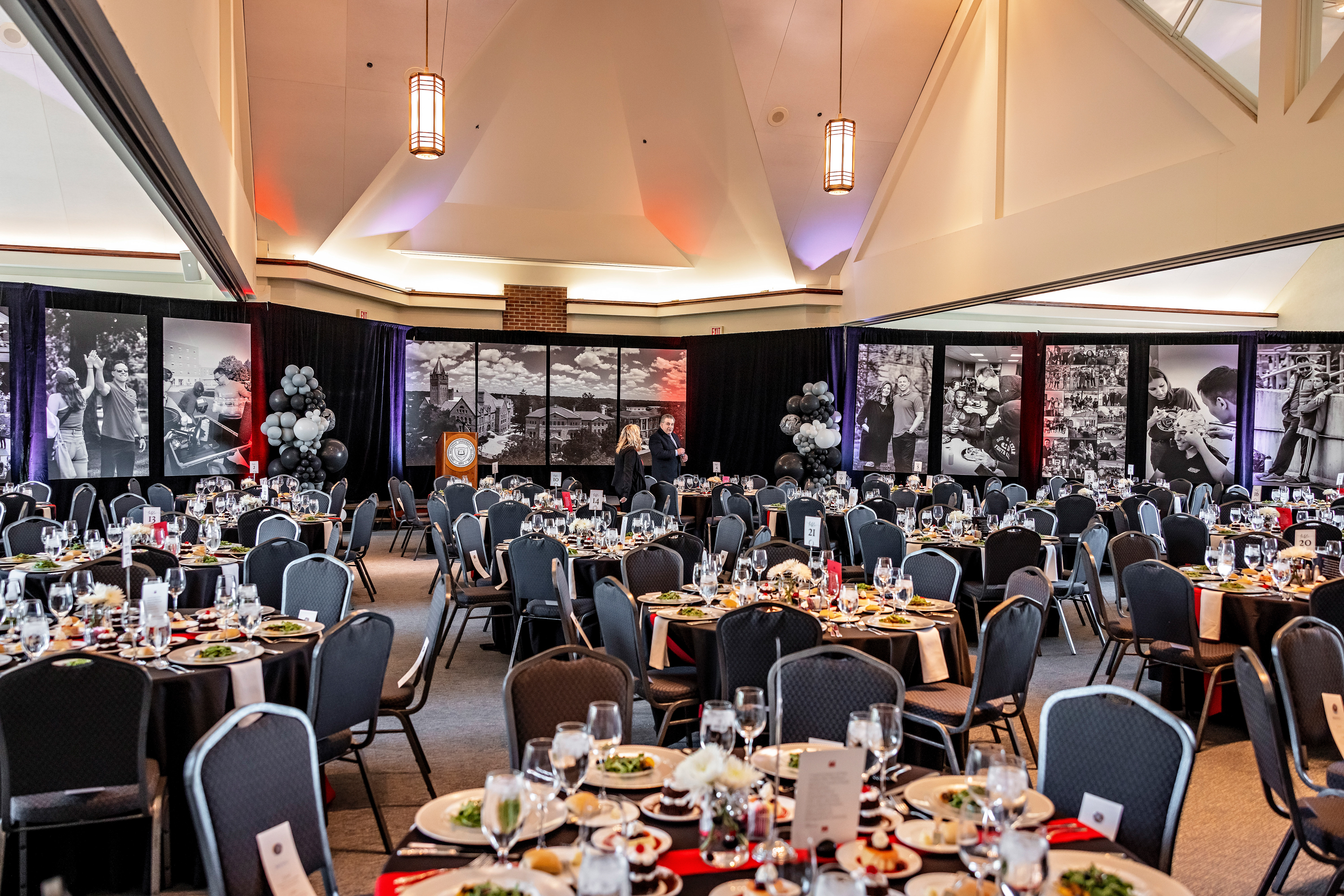 A ballroom is set up for a dinner event. Along the back wall, horizontal black and white photos hang and on the tables salad is served.