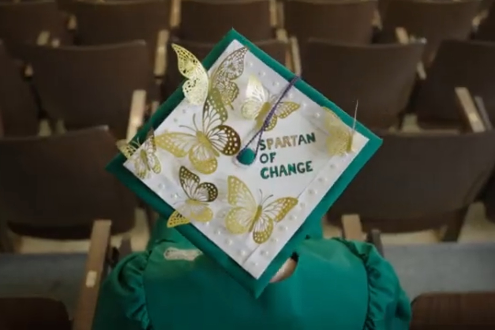 The top of a Michigan State graduation cap is decorated with gold foil butterflies and the words "Spartan of Change" in green foil.