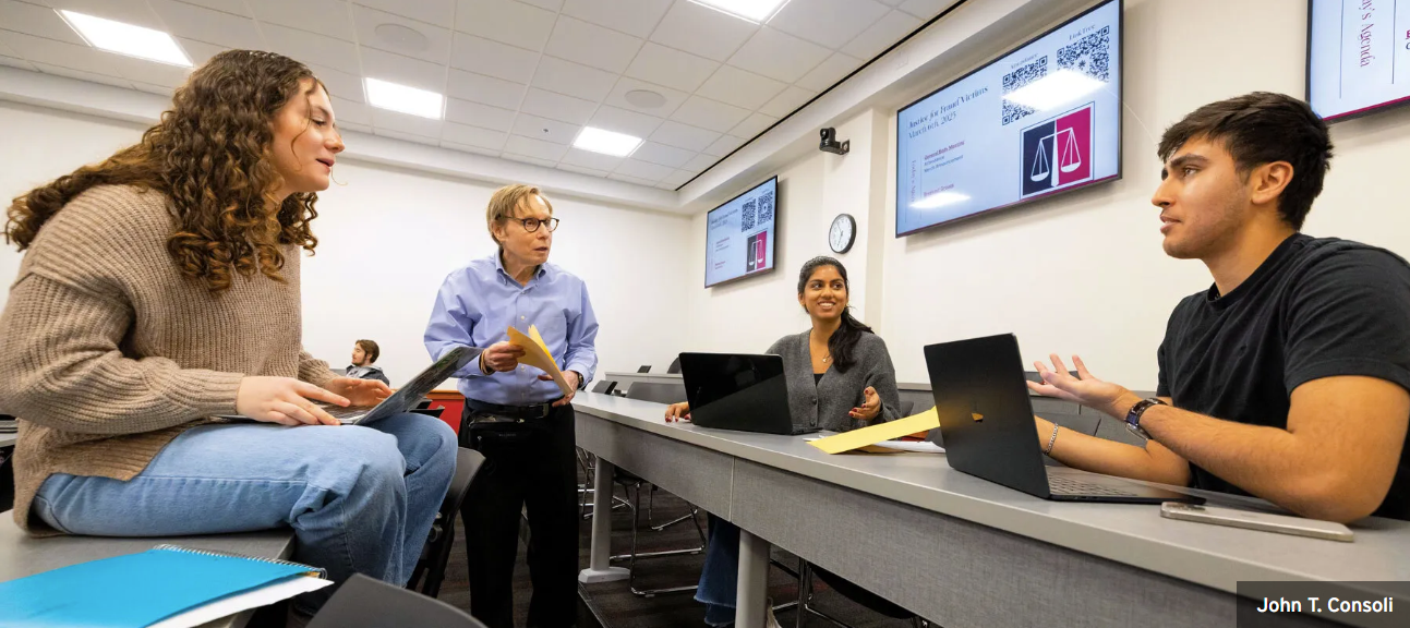 A photo of three students with laptops and a professor conferring in a classroom.