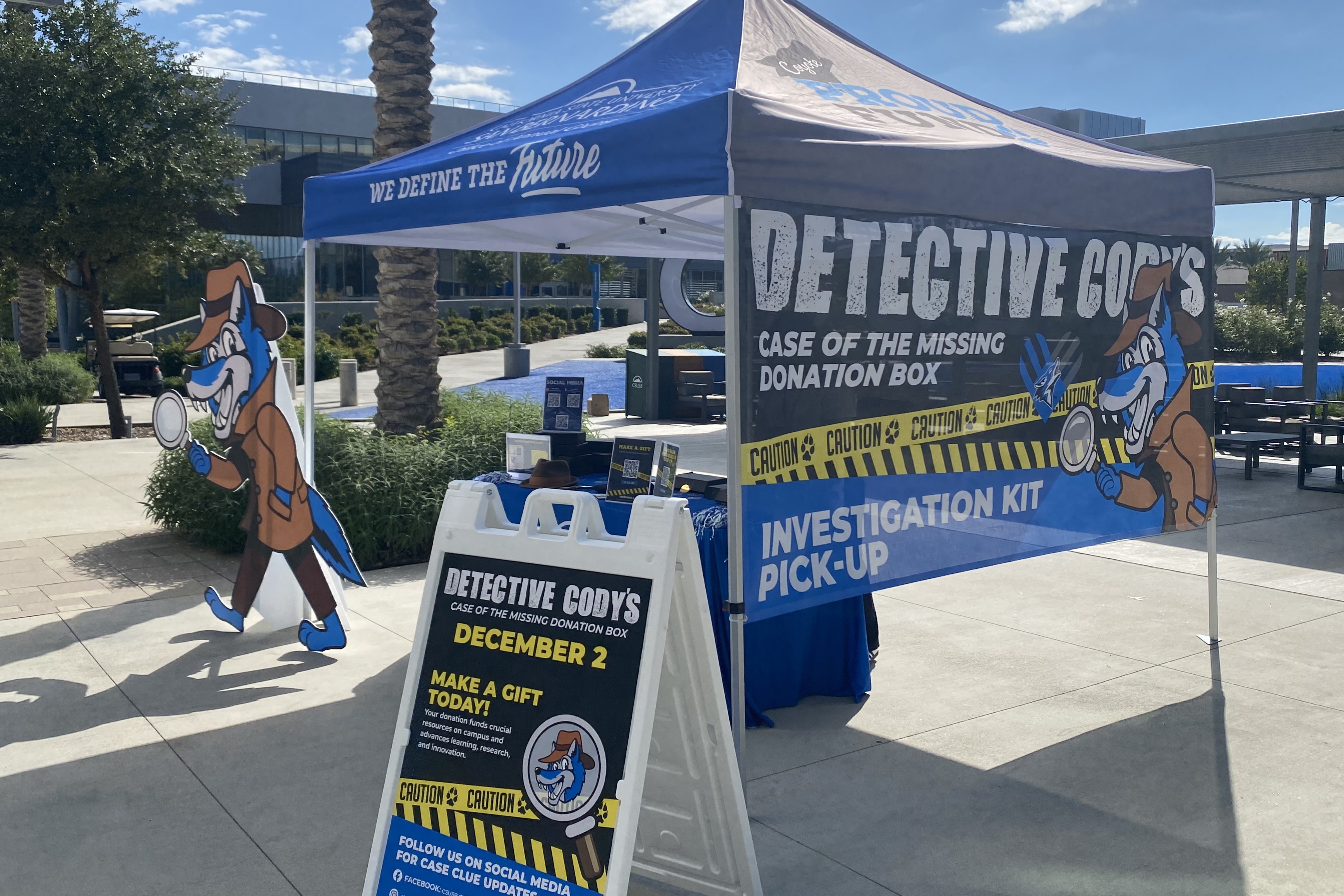 A photo of a blue tent set up for CSUSB Giving Day. Next to the tent is a cardboard cut out of the Detective Cody character, a blue wolf wearing a trench coat and a deerstalker hat and holding a magnifying glass.