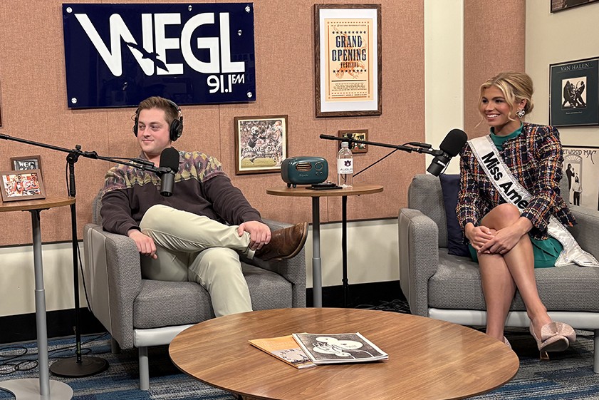 On the set of the "Everything Auburn" podcast, one of the hosts and a guest, a Miss America contestant, sit in chairs with microphones in front of them.