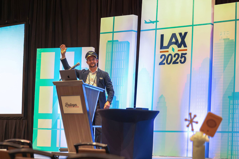 A photo of a man standing on stage at a podium wearing a sailor's hat. Behind him is a white and blue graphic with images of a skyline, a plane, and the text "LAX 2025."