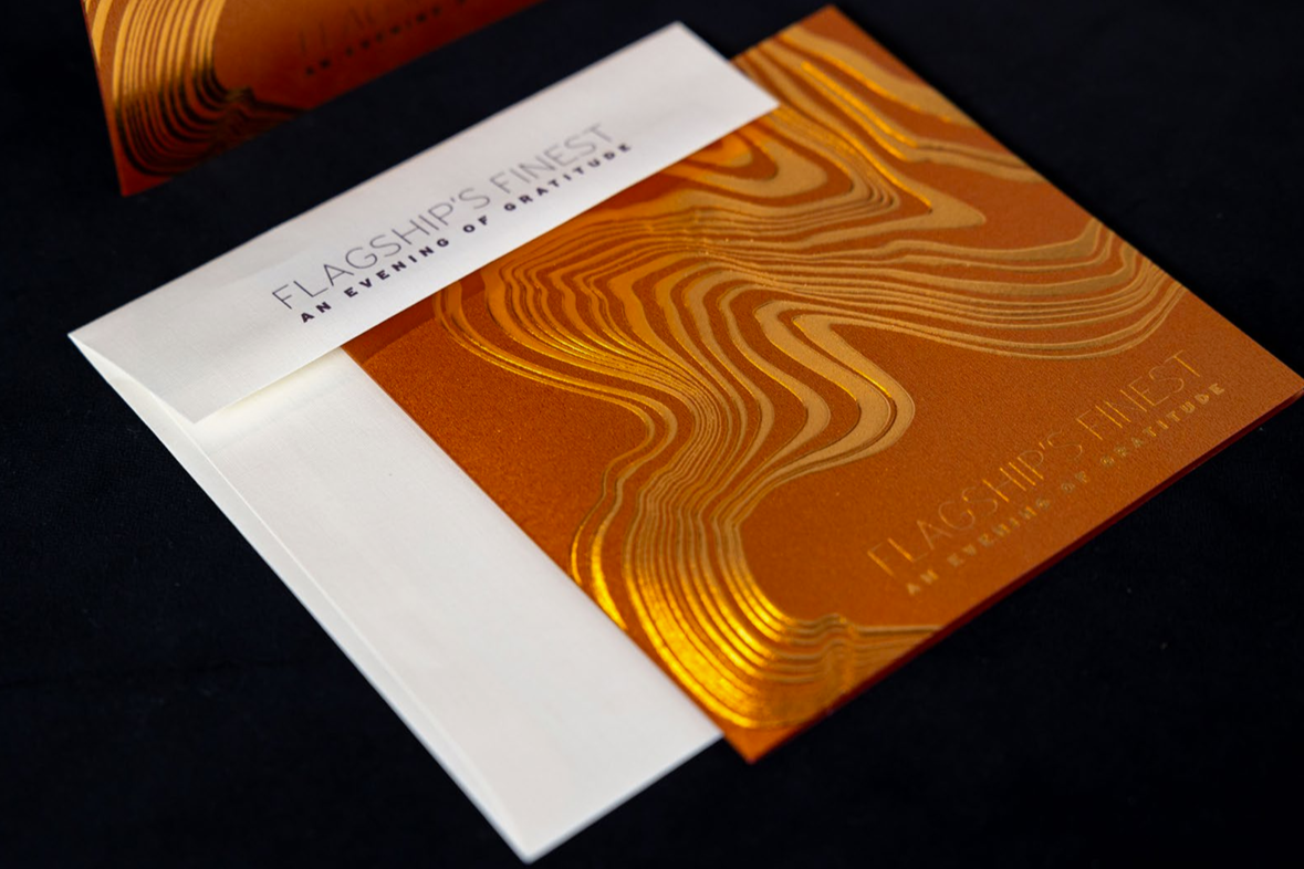 A photograph of University of Maryland's Flagship's Finest invitation. It's burnt orange in color with copper foil embossing and is paired with a white envelope.