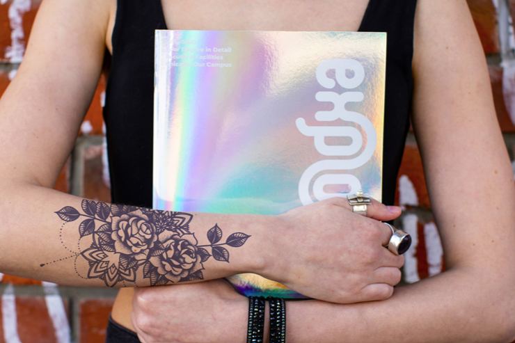Close up of someone holding SAIC's explore booklet in front of their chest. The shiny silver cover reflects in the sun.