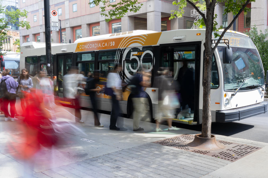 Photo of a shuttle bus with Concordia's 50th Anniversary-branded advertisement on its side. A line of people wait to enter the bus.