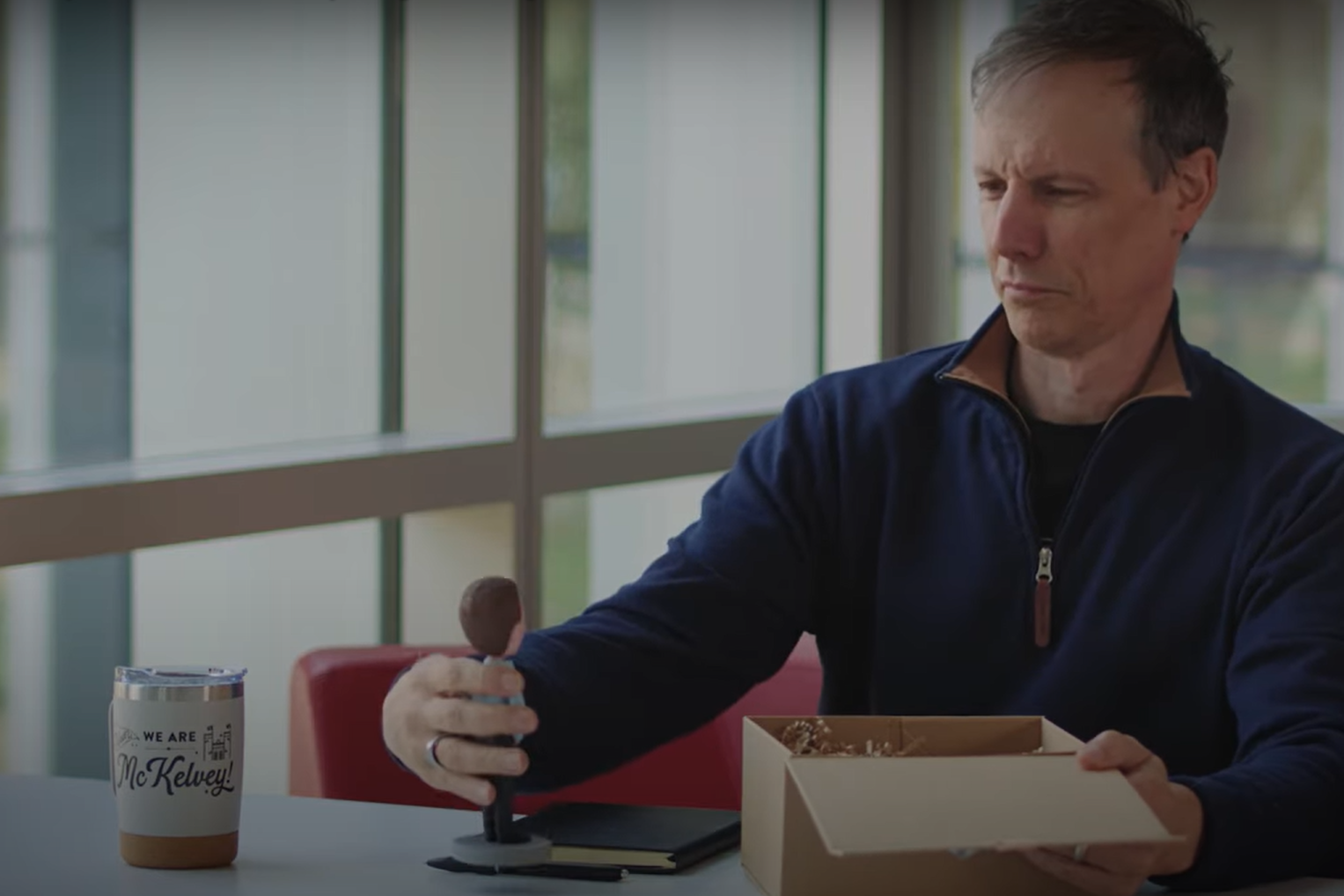 Sitting at a table, a man removes a bobble head of himself from a giftbox.