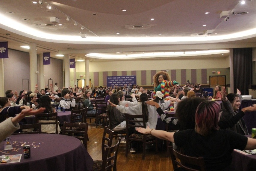 A ballroom decorated with K-State merch is full of alumni all looking to the drag queen standing in the center of the room