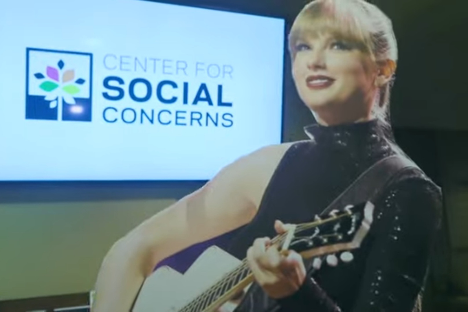 A cutout of Taylor Swift holding a guitar mid-performance is held up in front of a screen where the words "Center for Social Concerns" are displayed.