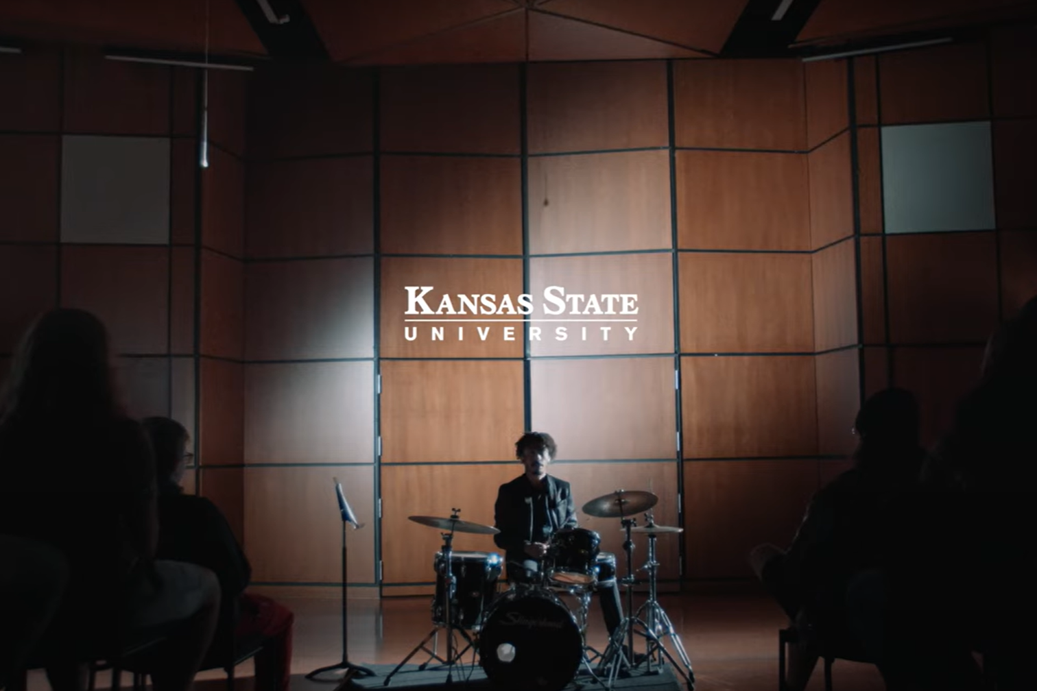 A drummer performs in a concert hall, his drum kit framed by a seated audience divided by a central aisle.