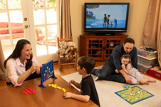 In a living room, a mother and son play Connect Four and a father and daughter play Chutes and Ladders.