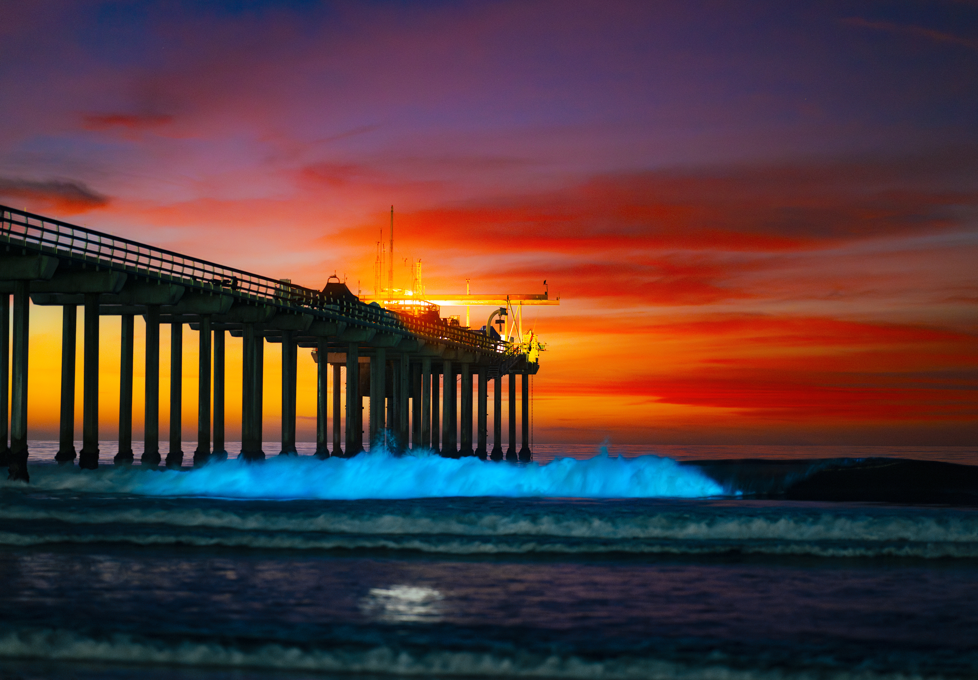 A pier enters the left side of frame. To its right, breaking waves glow neon blue. Behind it there is a red, yellow, pink and purple sunset.