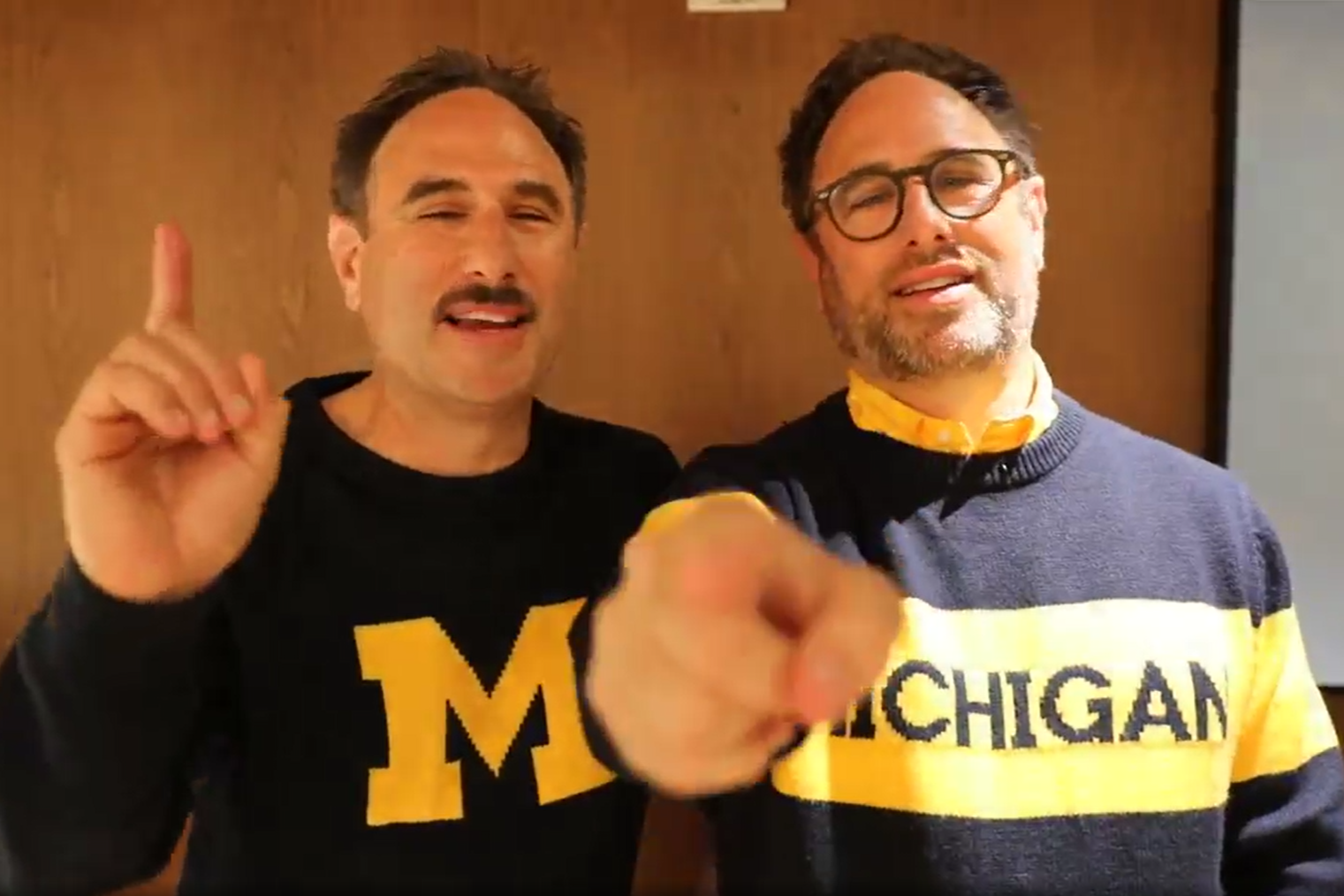 A screenshot from a video of the Sklar brothers. Both wear Michigan sweaters and point towards camera.