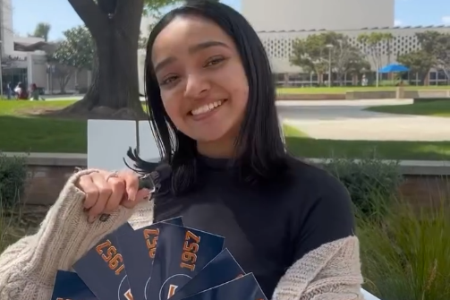 A Fullerton student holds a tiny mic in one hand and in the other holds Fullerton-branded stickers up to the camera.