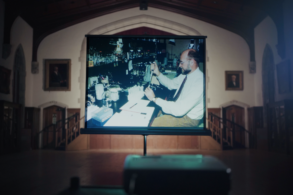 In a church, on a projector screen, an old photo of a man in a lab using a pipette.