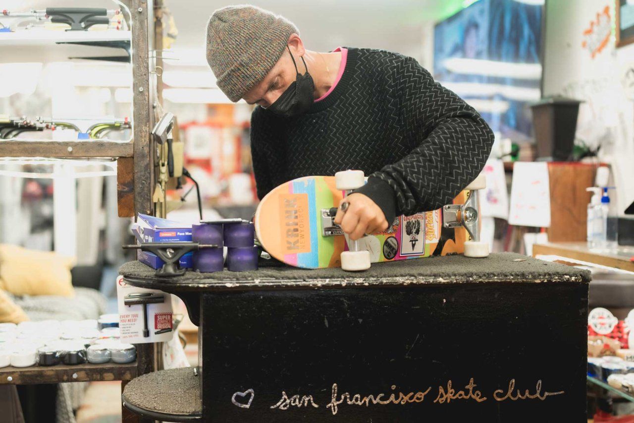 Shawn Connolly works on a multi-colored skateboard on a work table that has "San Francisco Skate Club" written on it.