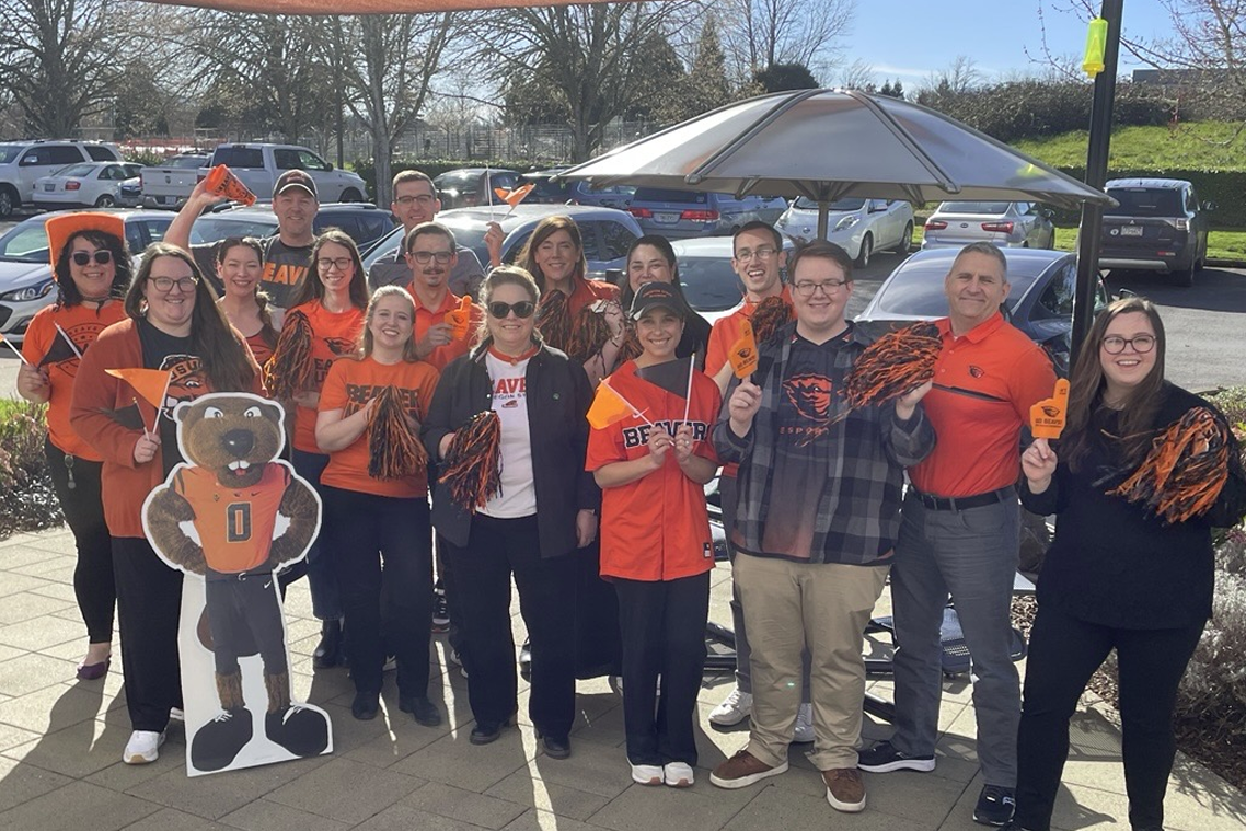 A group of people stand outside next to a parking lot wearing OSU-branded shirts and holding black- and orange-colored pom poms.