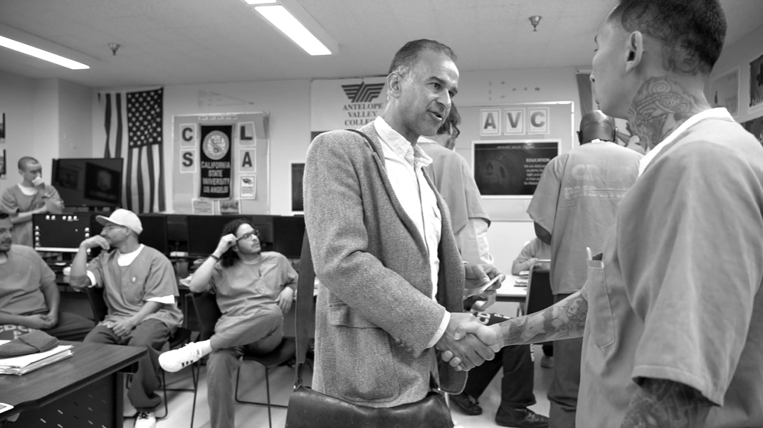 In a classroom, a tattooed man in a prison uniform shakes hands with a man wearing a suit coat and messenger bag.