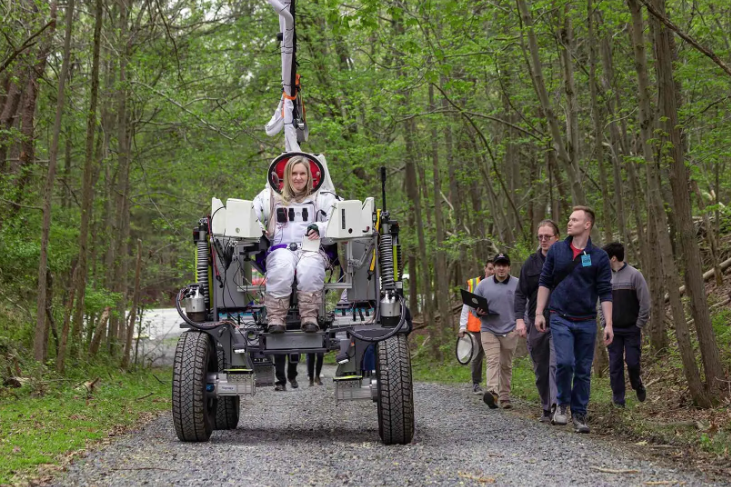 Maggie Haslam, dressed in a space suit, takes a rover prototype for a test drive through the woods.