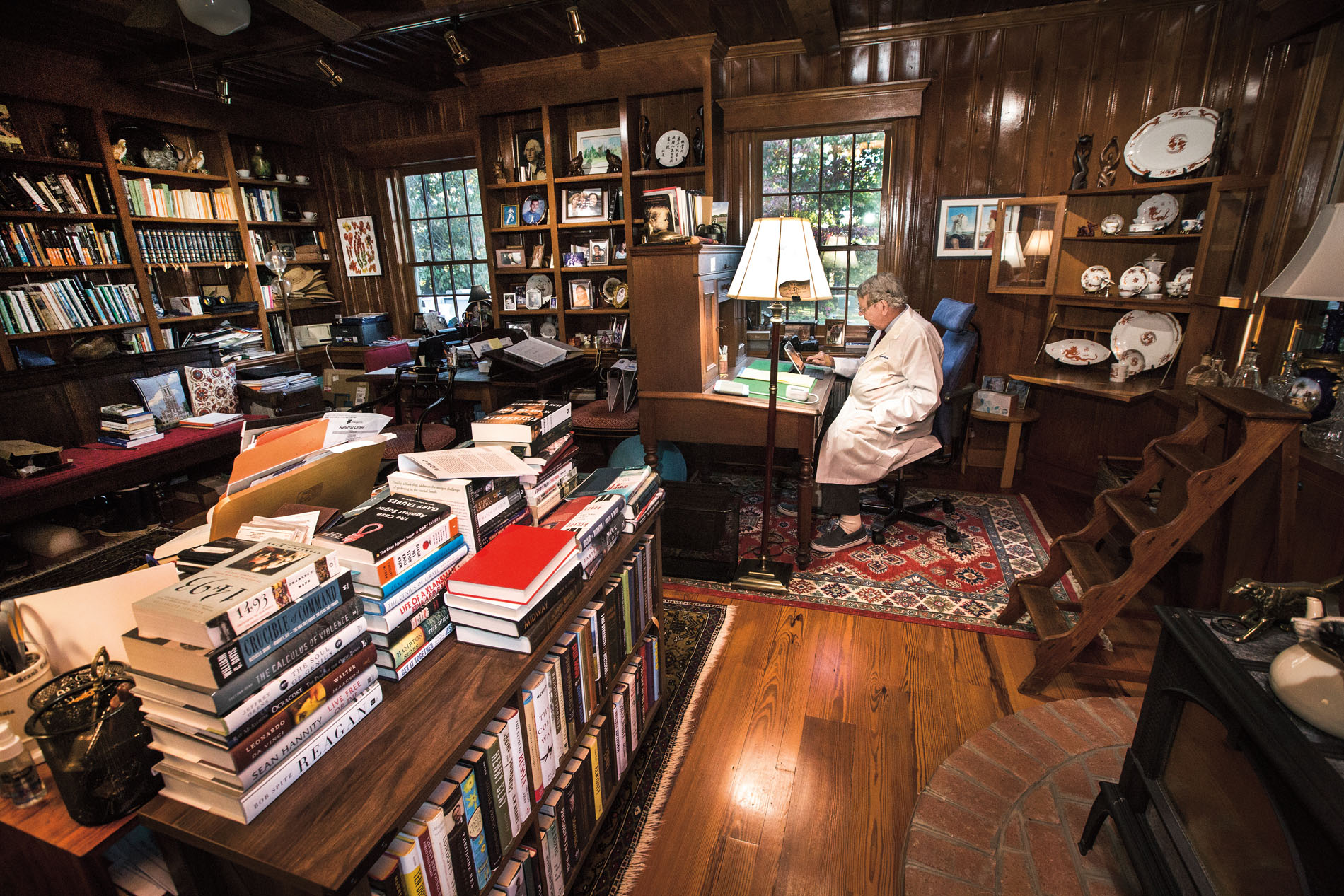 A Duke neuro-oncologist wearing a white coat sits in his office, which is filled with rugs, a table, couch, and selves filled with books, photo frames, dishware, and more.