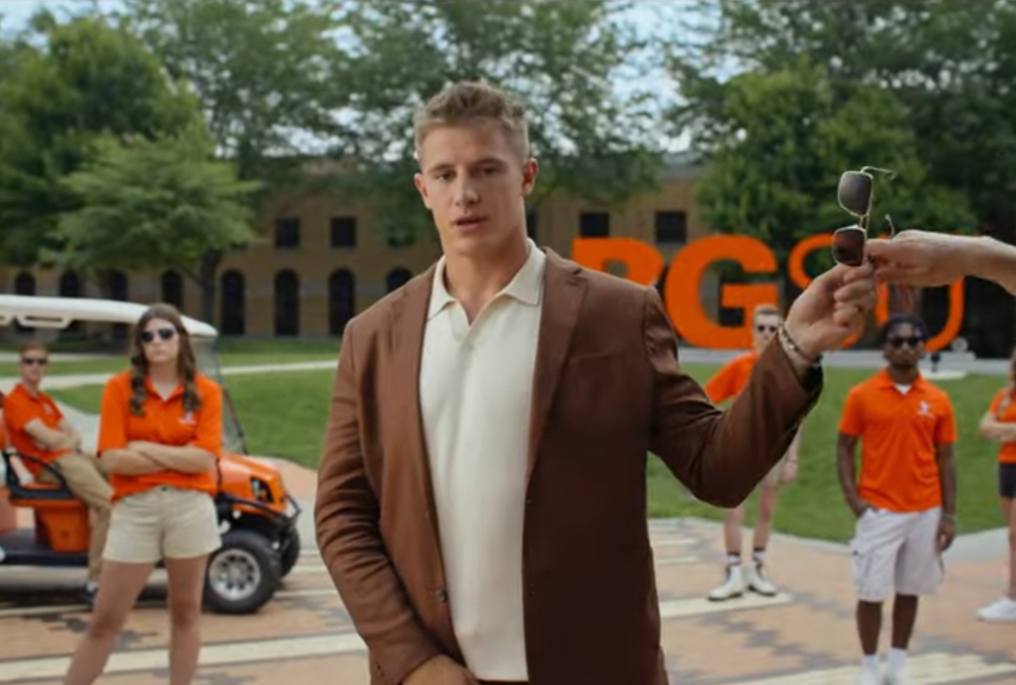A BGSU student hands his sunglasses to someone off screen. Behind him stand other students, all wearing matching orange polos.