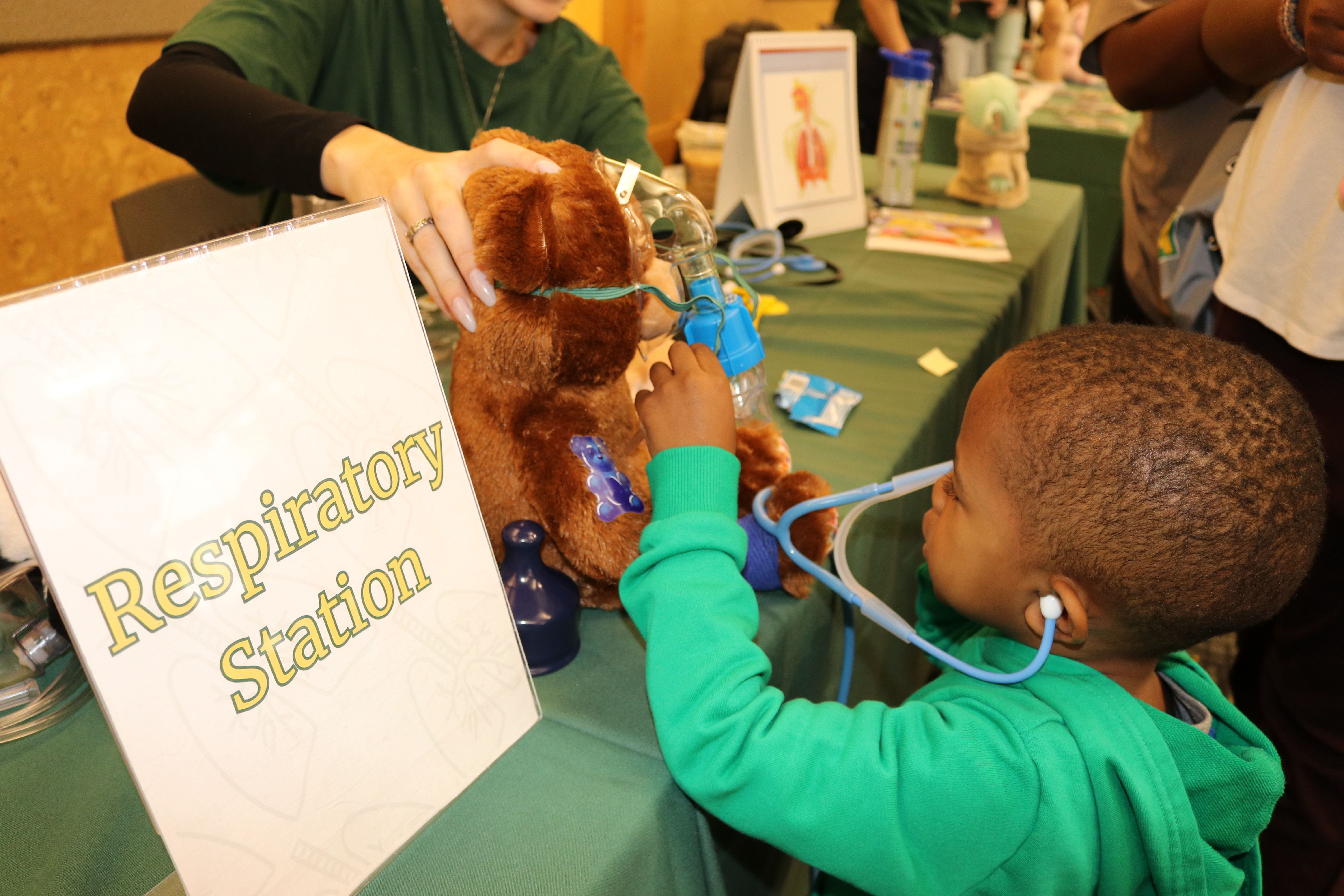 A child uses a toy stethoscope to listen to a stuffed bear's heart at a table with a "Respiratory Station" sign.