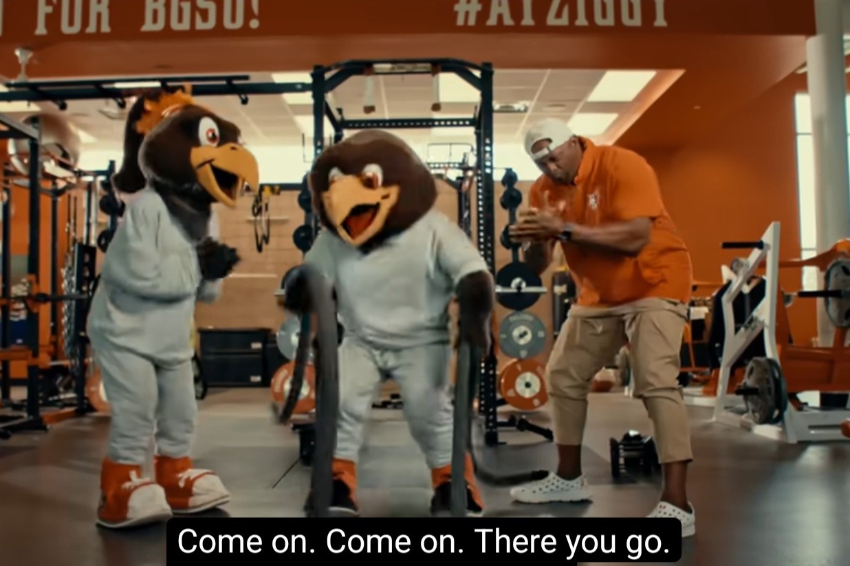 A screenshot from "Super Freddie." Two falcon mascots and a man train in a campus gym.