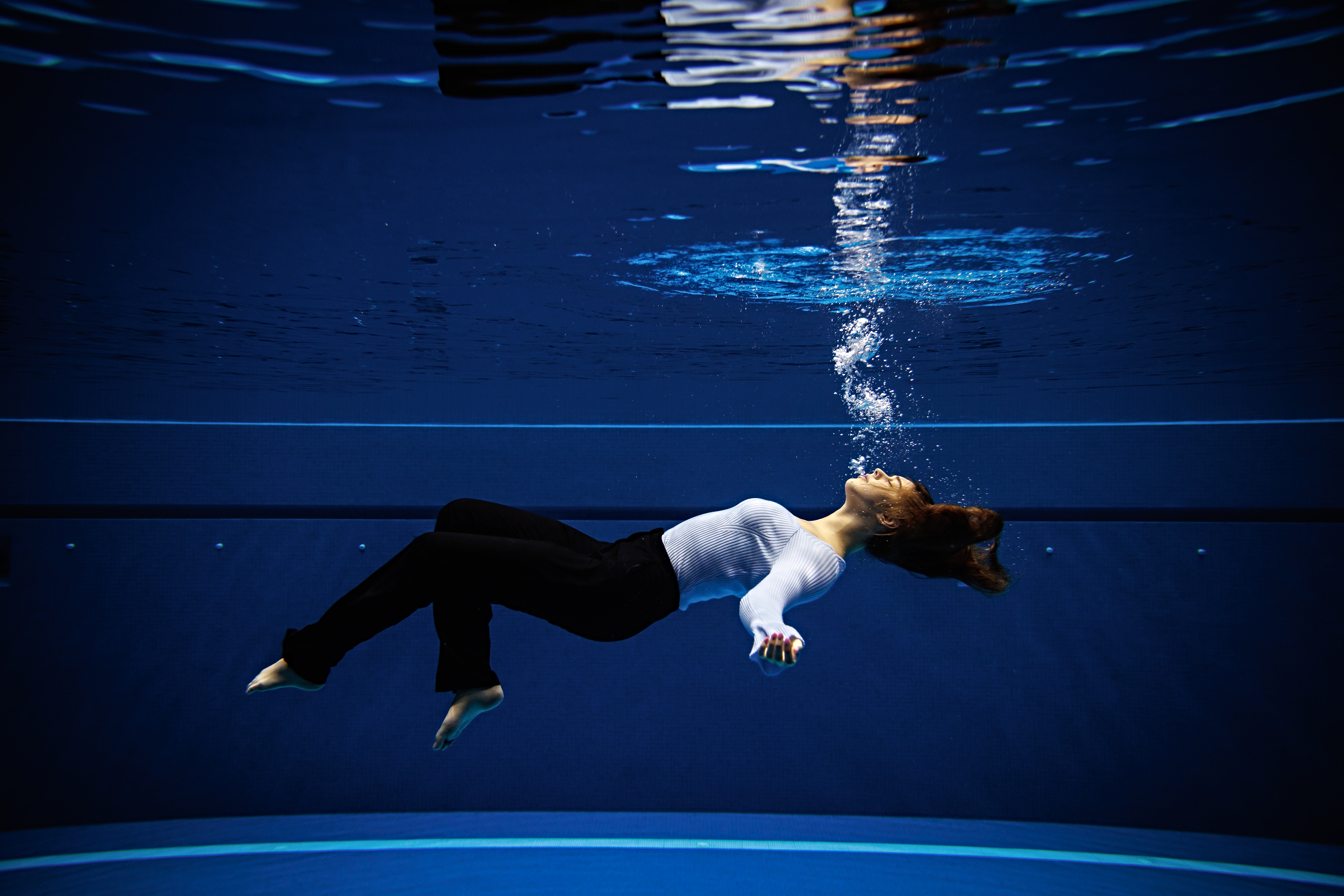 A girl dressed in business attire lays suspended and submerged in a pool. Air bubbles float from her mouth to the surface.