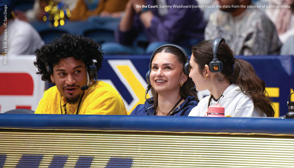 Three students sit courtside at a University of San Diego game wearing headsets.