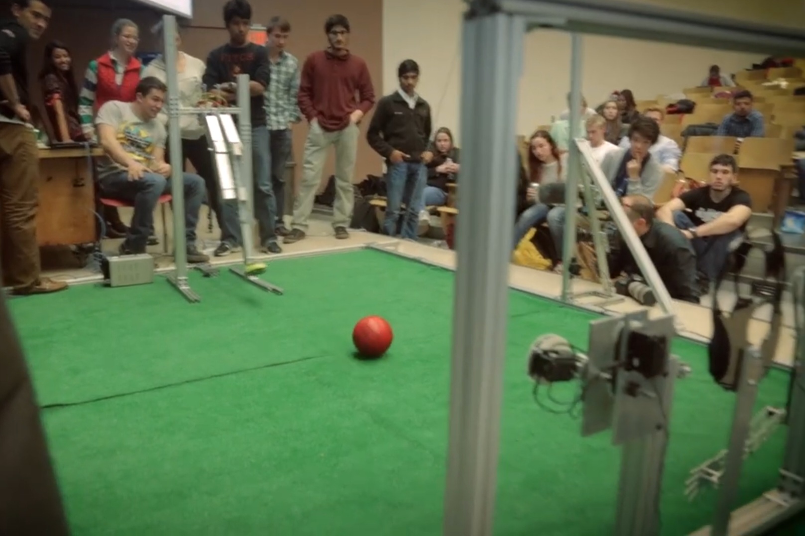 Students gather around a constructed field to watch mechanically engineered robots play with a red ball.