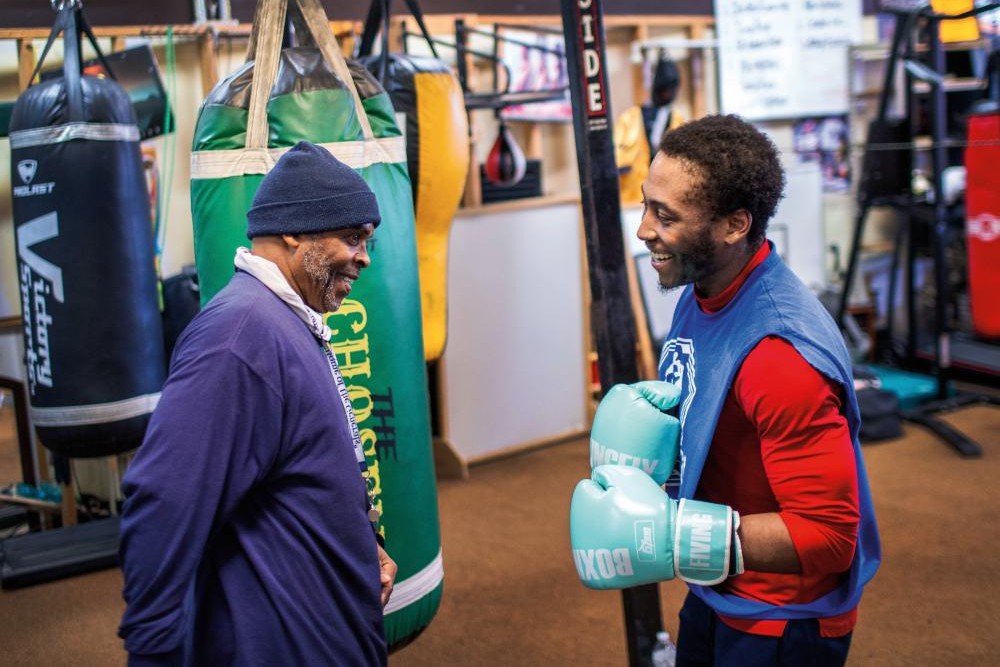 Walter Davis, wearing light blue boxing gloves, jokes with his coach Scott Brown in a boxing gym.