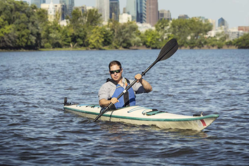 A man wearing a blue life vest kayaks in the middle of a river. In the background you can see a city skyline.