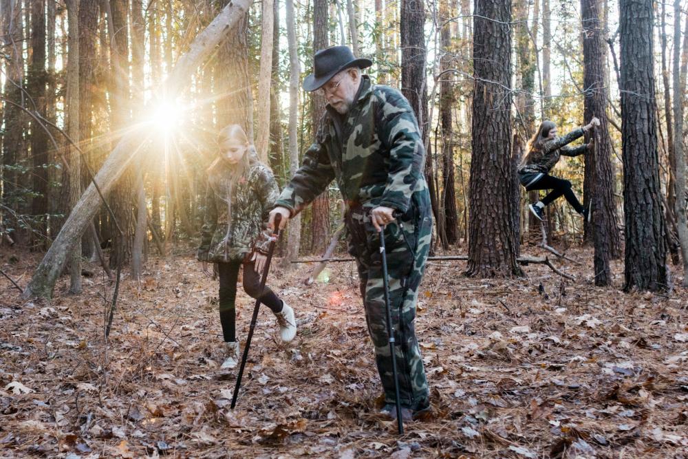 Bill Hicks uses two hiking sticks to walk on the forest's leaf-covered ground. Beside him walks a blonde girl, and another blonde girl climbs a tree truck in the background.