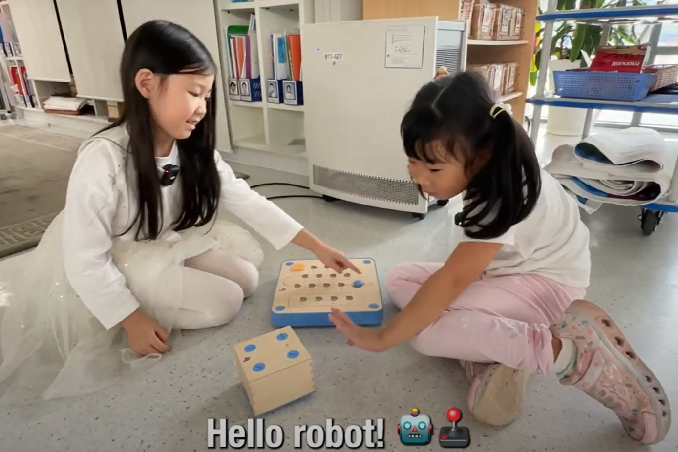 Two students in UNIS Hanoi's Early Childhood Centre sit on the ground with two block toys between them. One of them waves at the moving box, saying "Hi, Robot!"