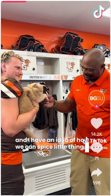 A screenshot from a BGSU TikTok shows a football coach shaking the paw of a cat being held by a football player.
