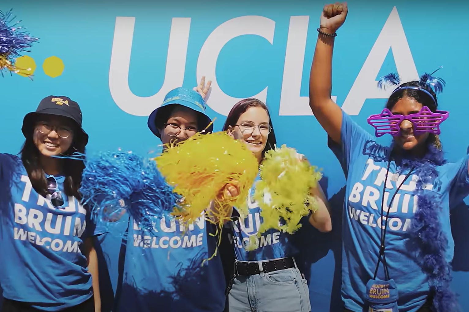 Four UCLA students pose for a photo. All are dressed in university merch. Three hold blue and yellow pom-poms and a fourth wears purple joke glasses and a blue feather boa.