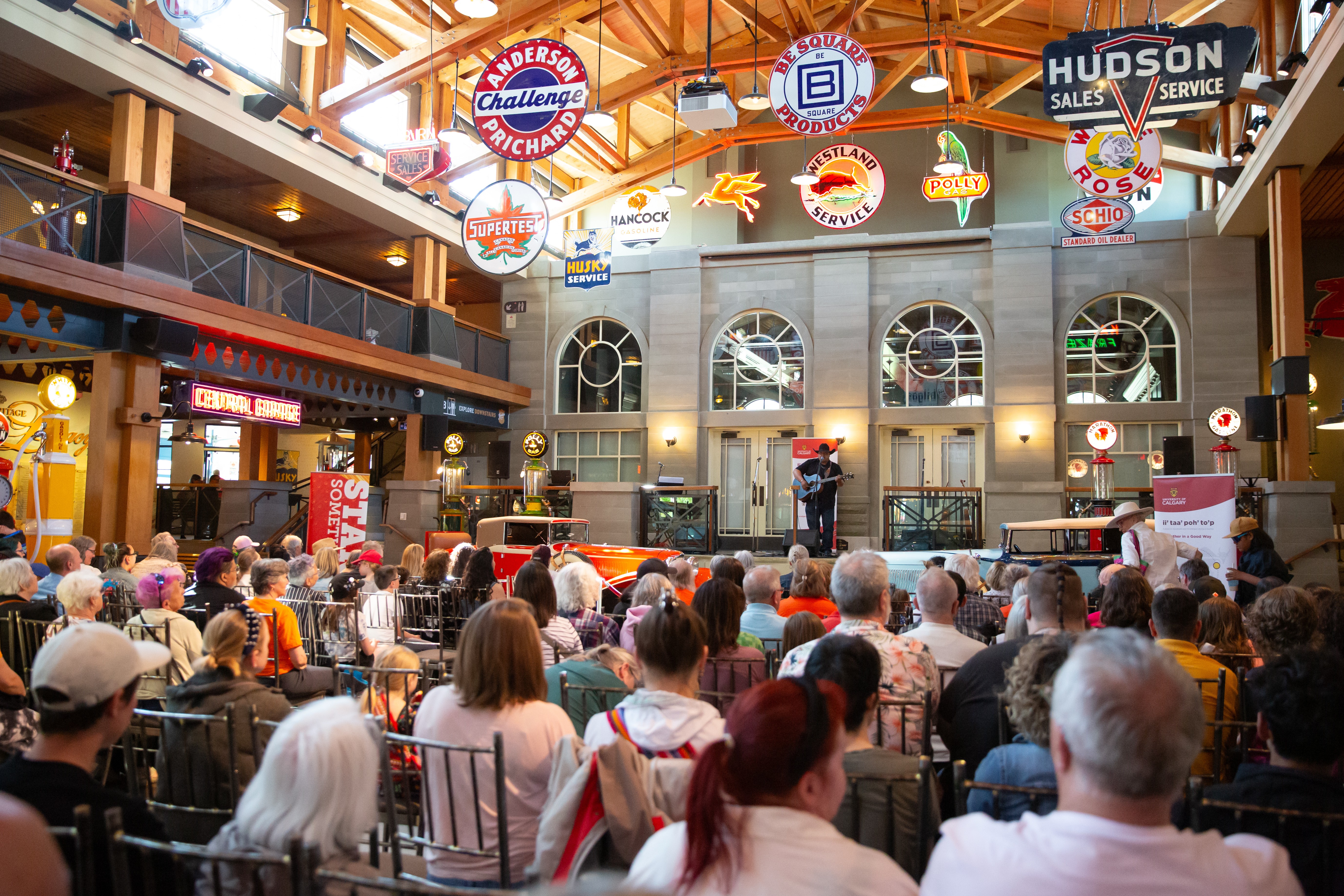 A seated audience watches a lone guitar player on stage. Vintage metal signs hang from the vaulted ceiling.