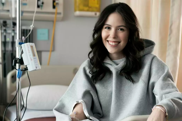 Maddison Tory smiles for a photo while sitting in a hospital bed wearing one of her IV-accessible hoodies.