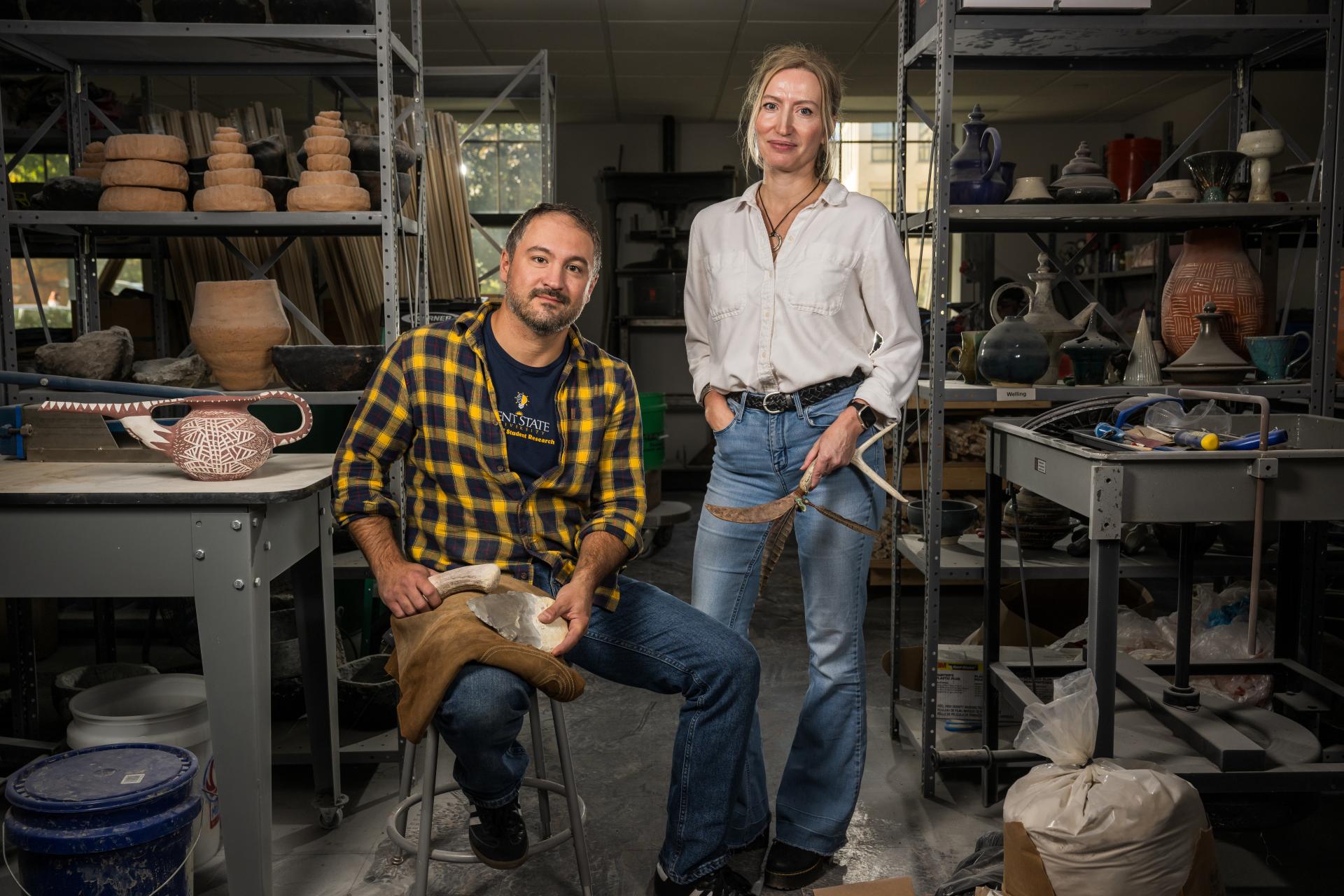 Metin Eren and Michelle Bebbers pose in a workshop filled with shelves of relics and archeology tools.