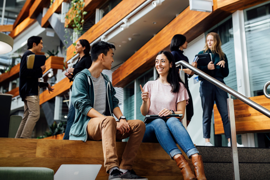 Two people sit on a staircase talking, while others move through the building behind them.